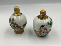 Two antique Chinese porcelain snuff bottles hand painted with traditional figures and nature scenes, gold-colored stoppers visible, placed next to a green apple for size reference.