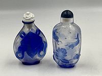 Two Asian snuff bottles made of blue and white glass, one with white stopper and abstract pattern, other with dark stopper and fish motifs.