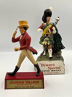 Photo shows both advertising figurines side by side, the Johnnie Walker figure in red coat on red base, Dewar's Special Highlander behind on rock base
