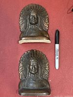 Pair of metal Indigenous Chief bookends with copper effect finish, detailed feathered headdress and facial features, placed on red fabric background next to a Sharpie marker for scale.