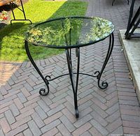 Full view of round cafe/bistro table showing clear glass top and curled black metal legs on brick patio.