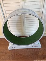Front view of circular cat treadmill exercise wheel with green turf inside, white frame, standing on wooden floor against white folding doors.