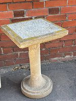 Front view of solid cement plant stand with square top and pedestal base against brick wall.