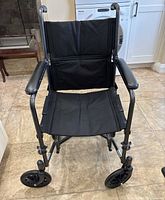 Front view of Drive transfer chair showing black fabric seat, cushioned armrests, and front wheels on tiled floor.