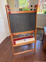Front side of the wooden child’s art easel displaying the black chalkboard surface and the unused roll of white Mala paper on the paper holder shelf.