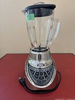 Oster blender front view showing glass jar, handle, black lid, chrome base with control buttons and power cord.