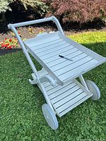 Photo showing the light gray outdoor serving cart from angled front view with top and bottom slatted shelves, curved handle, and two wheels on grass.