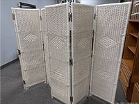 Two white wicker room dividers shown partially unfolded in a room with wooden furniture around. Each divider has four narrow, tall panels with diamond pattern in wicker weave.