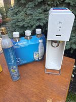 Photo showing the white Sodastream machine on a wooden surface outdoors, with the blue labeled CO2 cartridge and unopened pack of three Sodastream bottles beside it.