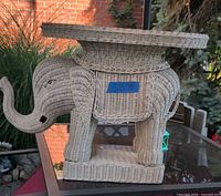 Side view of wicker elephant table showing trunk and woven body structure, outdoors on glass table.
