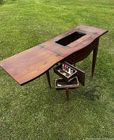Photo of antique solid wood sewing table unfolded on grass showing one fold-out side leaf extended with pull-out drawer open holding sewing spools and tools.