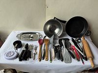 Full view of all kitchen utensils and cookware spread on a white cloth showing metal cooking pot with lid, cast iron pan, oval tray, salad bowl, rolling pin, measuring cups and various utensils mainly black, wooden and metal.