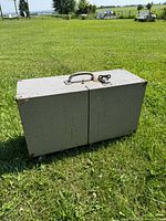 Closed toolbox with visible handle on top and vinyl-coated wood surface. The toolbox is rectangular with metal corners and sits on grass outdoors.