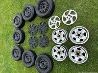 Photo showing assorted black steel rims mounted with tires, various sizes, placed on grass with hubcaps and other rims nearby