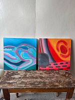 Pair of abstract acrylic paintings on canvas placed side by side on top of a distressed wooden table against a plain wall, showing front views of both paintings with visible brushstrokes and colors.