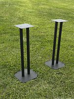 Pair of black matte speaker stands shown outdoors on green grass facing forward, showing top speaker plates and cylindrical supports.