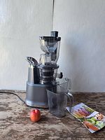 Side view of SiFENE cold press juicer with juice and pulp containers attached on wooden surface. Clear plastic components and chute visible. Single apple placed for scale. Original box in background partially visible.