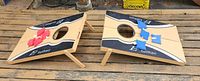 Two wooden bean toss boards with red and blue bean bags, legs unfolded, placed on wooden pallet.