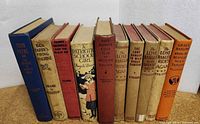 Ten vintage children's books arranged standing side by side, showing worn cloth hardcovers mostly beige and blue with varying author names and titles.