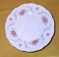 Single white china side plate with pink roses, blue accents, silver scroll work, and scalloped edges with silver trim on a yellow background.