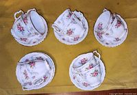 Top view of arranged sets of five tea cups with matching saucers, set on a yellow cloth, showing floral patterns and handles.