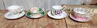 Four china tea cups with saucers displaying various floral designs lined up on a wooden surface.