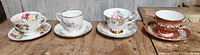 All four china tea cups placed in a row with matching saucers, showing variety of floral and gold patterns.