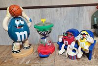All three M&M collectible figures shown together: blue basketball player, vintage spiral dispenser, red and yellow sitting figures with popcorn.