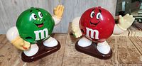 Front view of green and red M&M's collectible figurines on brown bases, showing waving hands and smiling faces.