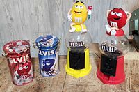 Four M&M collectible items lined up: two Elvis character tins on the left and two character dispensers on the right