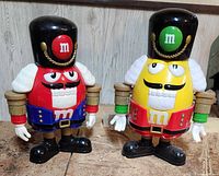 Photo showing the red and yellow M&M nutcracker collectible figures standing side by side on a textured surface with a wooden background.