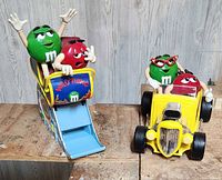 Both M&M collectible figurines with characters in vehicles: one in a roller coaster, the other in a hot rod car.