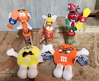 Five M&M's candy dispensers topped with character figurines holding fans and two M&M's plush toys