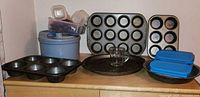 Photo showing all items together on a wooden surface: three muffin tins (12-hole regular, 6-hole regular, 6-hole extra large), salad spinner, one-cup glass measuring cup, lunch storage container, tin pie plate, pizza pan, and extra small storage containers.