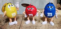 Photo showing the full view of 3 M&M standing collectible figures in yellow, red, and blue with two original tags attached.
