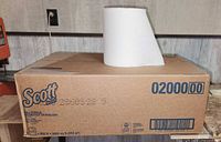 Photo showing full unopened brown cardboard box labeled with Scott brand and six paper towel rolls. A single sample white paper towel roll is sitting on top of the box displaying texture and size.