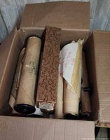 Open cardboard box containing several large piano player rolls, some rolled paper and some in decorative cardboard sleeves, different sizes and conditions.
