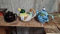 Lot of three tea pots shown on a rustic wooden surface against a gray wood paneled background