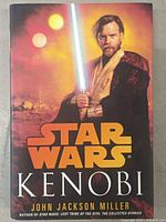 Front cover of "Star Wars Kenobi" hardcover book showing Obi-Wan Kenobi holding a glowing lightsaber with twin suns and a desert city background.