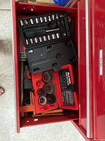 Top view of a red plastic toolbox drawer containing multiple socket wrenches and a black case with small sockets, along with a red plastic hole saw set in its labeled case.