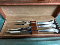 Set of three stainless steel carving utensils laid in wooden velvet-lined box, showing arrangement and details of knife blades and fork tines.