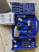 Full open blue molded plastic case showing air tools and accessories arranged inside. Three main air tools visible are an impact wrench, a ratchet, and an air hammer. Various sockets, bits, adapters, and fittings are organized in molded compartments. Instruction manual and packaging also present.