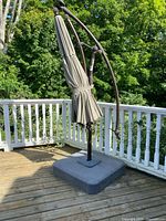Full view of cantilever patio umbrella with beige canopy and brown metal frame mounted on gray weighted base on wooden deck