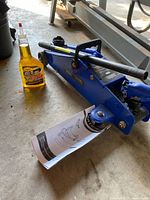 Blue Certified brand trolley jack with attached handle and manual underneath, alongside bottle of RisLone jack oil on concrete floor.