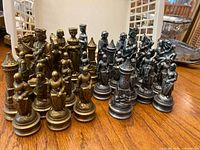 Set of bronze and silver colored metal chess pieces arranged on a wooden table with visible wear and missing pawns.