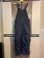 Full black insulated bib overalls hanging against a wooden background, showing overall length and structure.