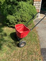 Side angle view of red broadcast push spreader showing plastic hopper and black handle