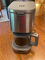 Full coffee maker unit showing stainless steel body, glass carafe with handle, and lid.