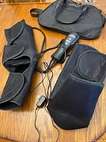 Full view of black fabric leg massager wraps, remote controller with attached wire, power adapter, and Fitpulse carrying bag on wooden floor.