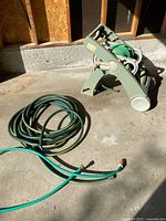 Garden hose attached to plastic wheeled reel cart, displayed unfolded on concrete floor with bright sunlight casting shadows. Hose is coiled and the reel cart is standing upright with wheels visible.
